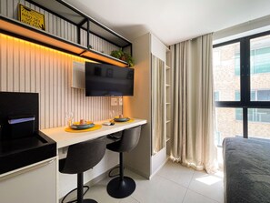 A modern hotel room with a flat-screen TV mounted on the wall, a dining area with a table and chairs, and a large window with curtains. - Get One By Hosppedar (João Pessoa)