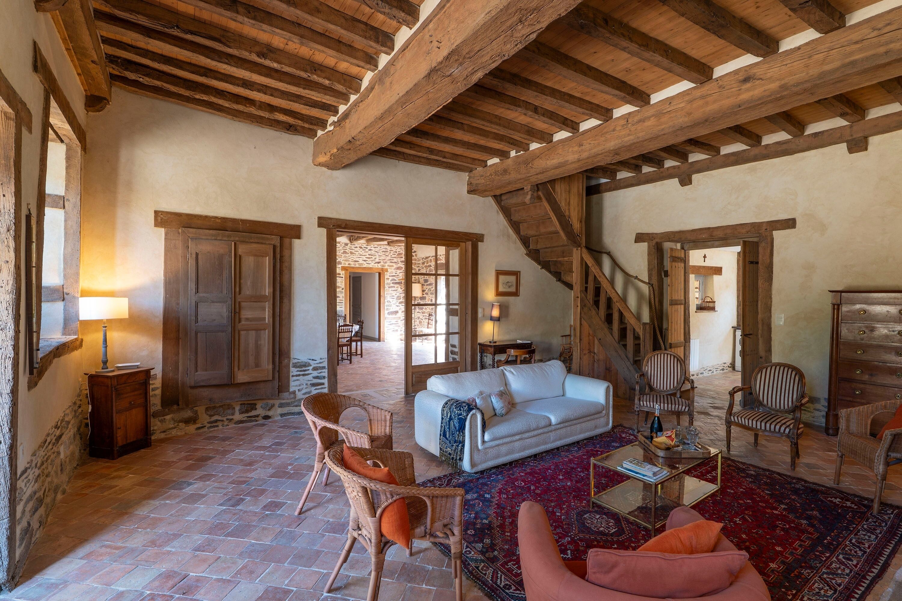 A rustic interior with exposed wooden beams, a white sofa, wicker chairs, and a patterned rug.