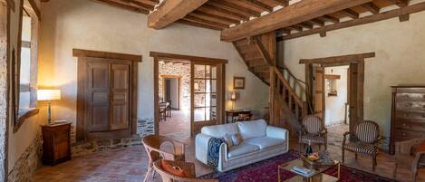 A rustic interior with exposed wooden beams, a white sofa, wicker chairs, and a patterned rug.