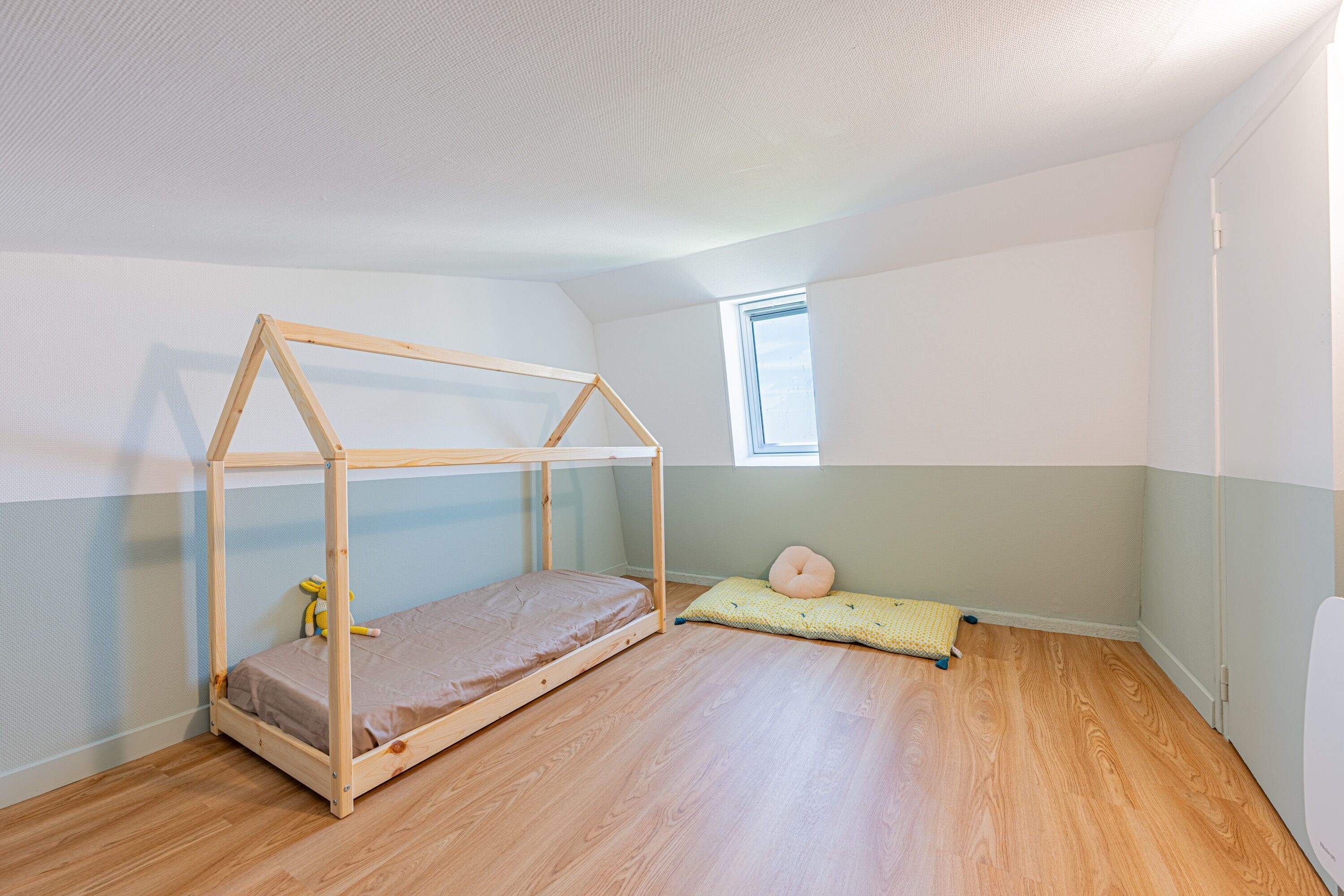 Ein Zimmer mit einem hölzernen Etagenbett, einem kleinen Fenster und einem grünen Kissen auf dem Boden.