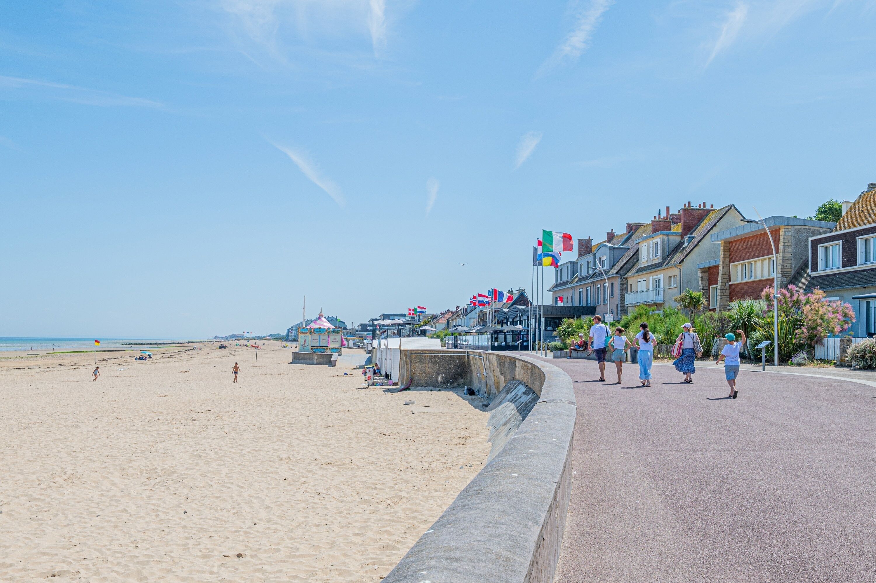 Une promenade en bord de mer avec une plage de sable, une digue en pierre incurvée et une rangée de bâtiments ornés de drapeaux.