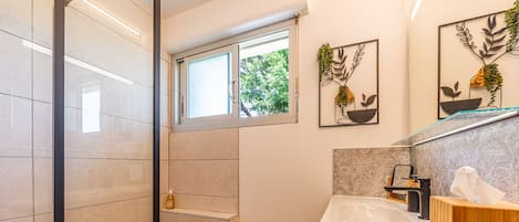 A modern bathroom with a glass shower enclosure, a wooden floor, a white sink, and a window with a view of greenery.