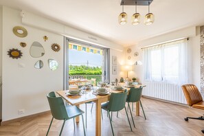 A dining area with a table set for four, a view of greenery outside, and a window with curtains. - L'Écho des Marées - Bernières-sur-Mer (Bernières-sur-Mer)