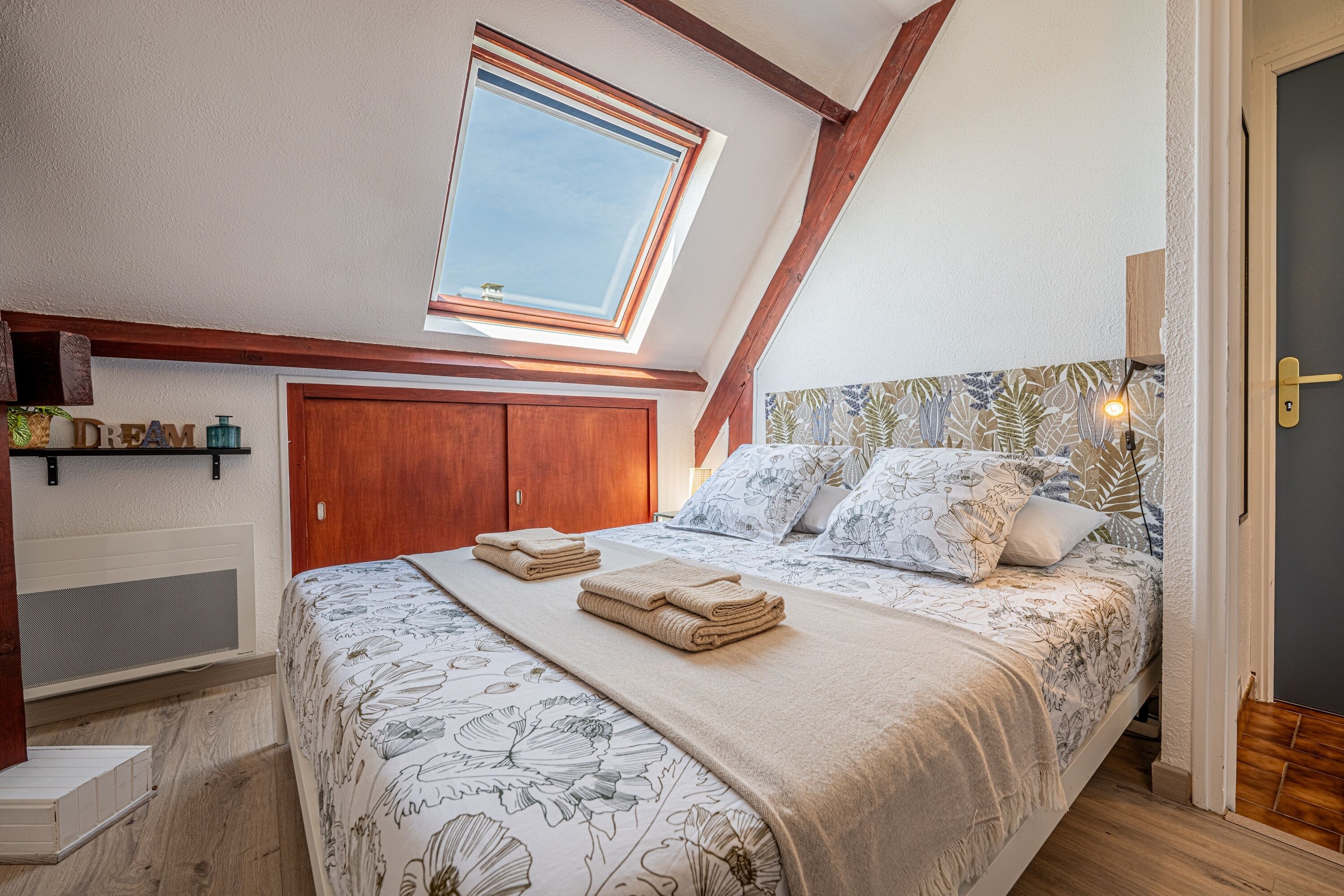 Un dormitorio con techo inclinado, tragaluz, cama con ropa de cama floral, armario de madera y mesita de noche con lámpara.