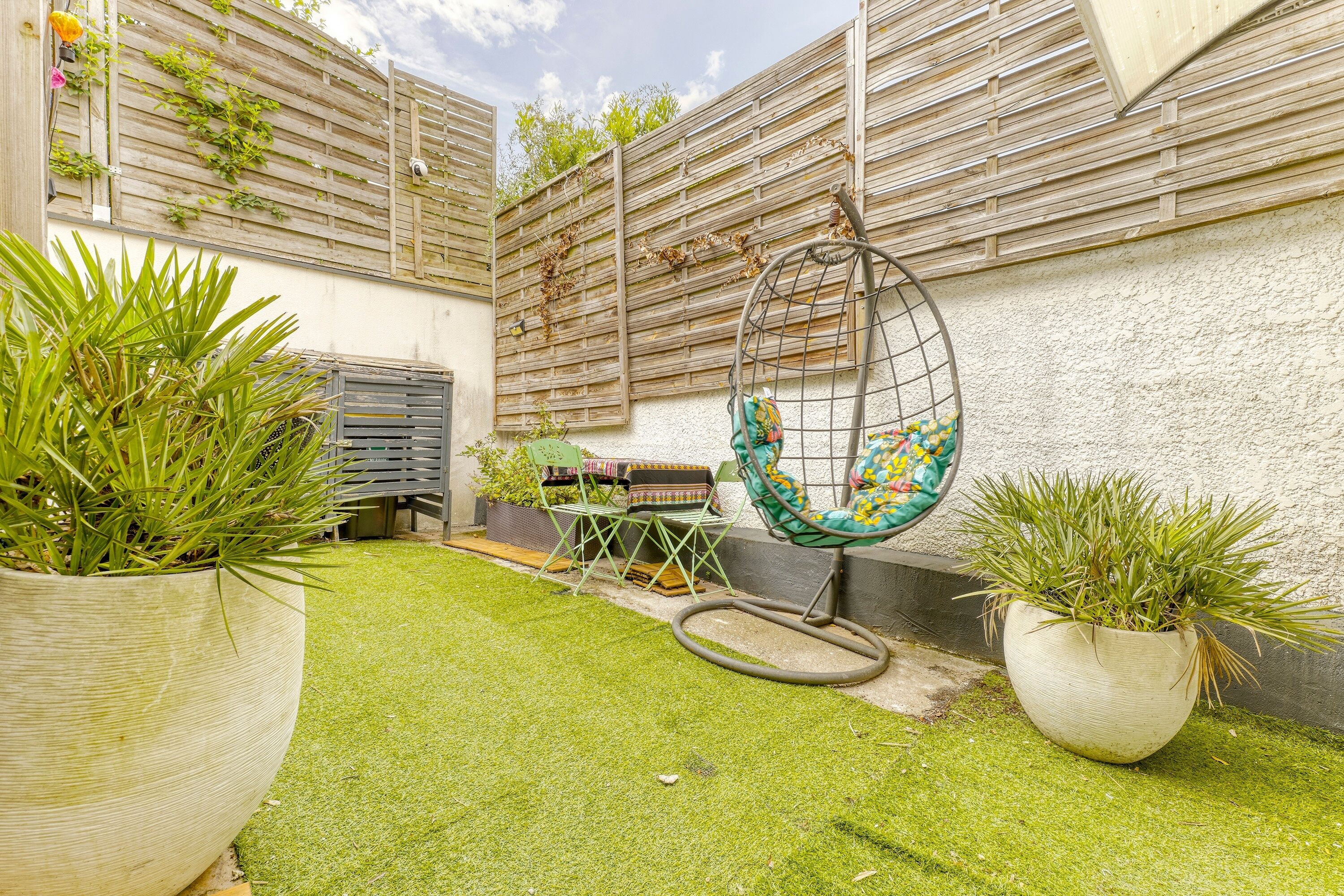 Un petit jardin clos, doté d’une pelouse artificielle, de plantes en pot, d’une chaise suspendue et d’une petite table avec une chaise.