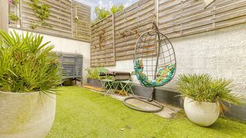Un petit jardin clos, doté d’une pelouse artificielle, de plantes en pot, d’une chaise suspendue et d’une petite table avec une chaise.