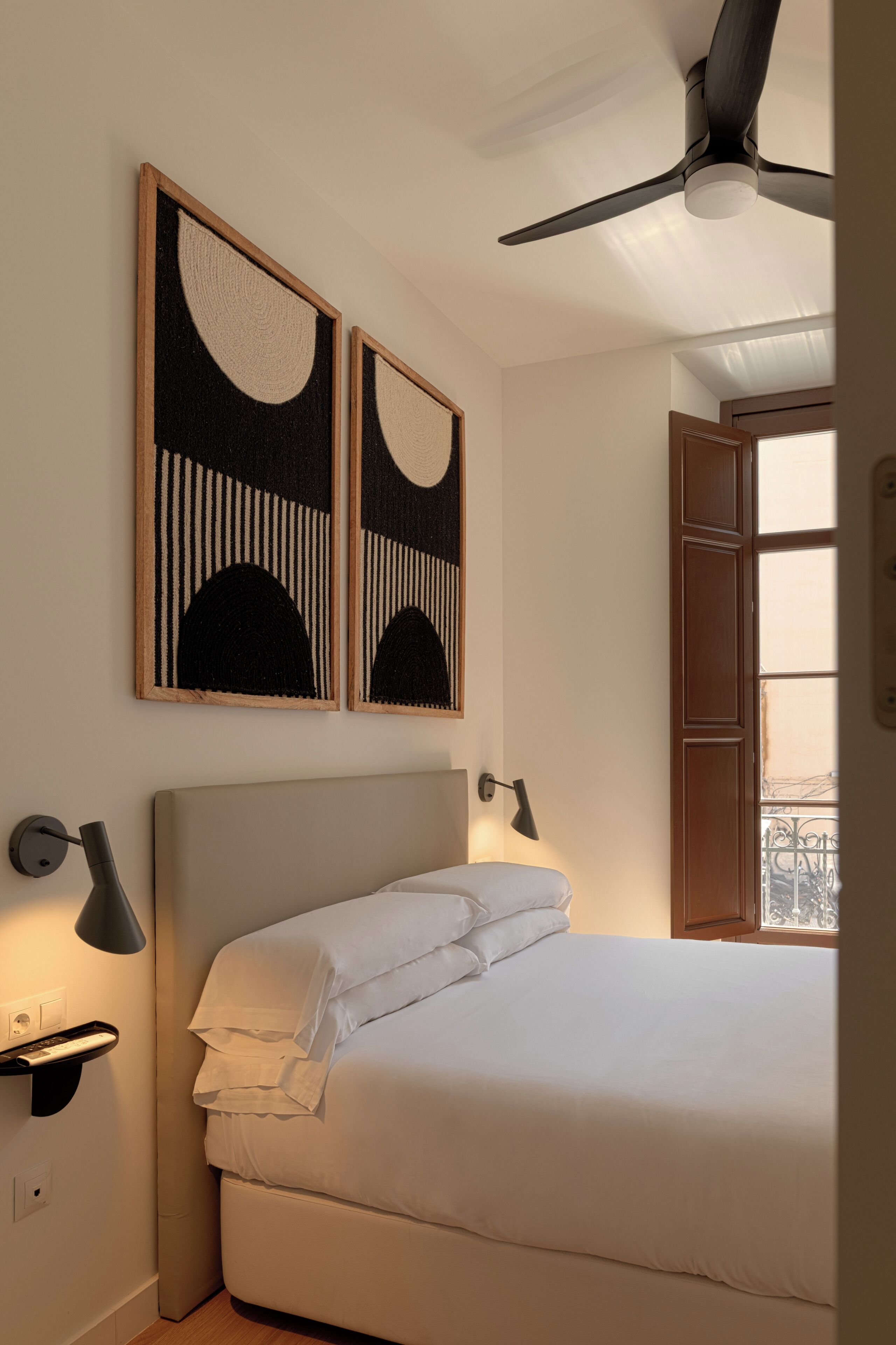 A bedroom with a bed, two wall-mounted lamps, a ceiling fan, and two framed artworks.
