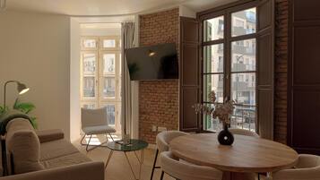 Een moderne woonkamer met een ronde houten tafel, twee fauteuils, een bank, een tv die aan een bakstenen muur bevestigd is, en een uitzicht op de stad door grote ramen.