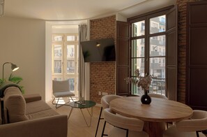 A modern living room with a round wooden table, two armchairs, a sofa, a TV mounted on a brick wall, and a view of the city through large windows. - El Racimo Apartments (Málaga Capital)