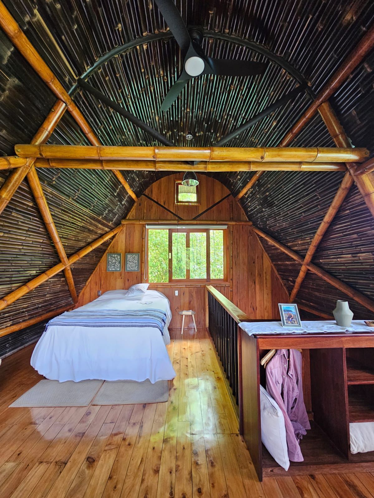 A room with a sloped ceiling, wooden beams, a bed with a white bedspread, a small table with a vase, and a window with a view of greenery.
