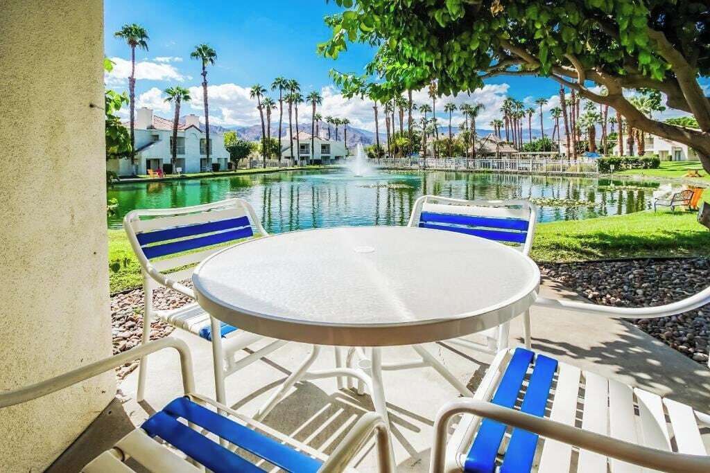 A round outdoor table with blue and white chairs, a pool with a fountain, and palm trees in the background.