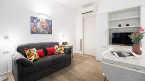 A modern living room with a grey sofa, colorful cushions, a painting on the wall, a white door, and a kitchen area with a TV and a potted plant. - Italianway Comfort - Bligny 19 E (Milan)