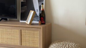 Eine Holzkonsole mit einem gerahmten Bild von Surfbrettern, einer Champagnerflasche und einem gestrickten Pouf vor einer Wand.