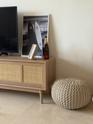 Eine Holzkonsole mit einem gerahmten Bild von Surfbrettern, einer Champagnerflasche und einem gestrickten Pouf vor einer Wand.