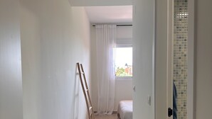 Ein schmaler Flur mit einer Holzleiter, die an der Wand lehnt und zu einem Zimmer mit einem Bett und einem Fenster führt.