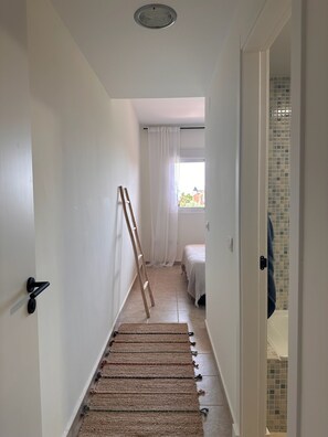 Ein schmaler Flur mit einer Holzleiter, die an der Wand lehnt und zu einem Zimmer mit einem Bett und einem Fenster führt.