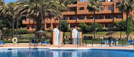 Een hotel met een zwembad, palmbomen en een helderblauwe lucht.
