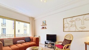 Ein Wohnzimmer mit einer Couch, einem Fernseher und einem Sessel. Ein Fenster bietet einen Blick auf die Gebäude außerhalb.