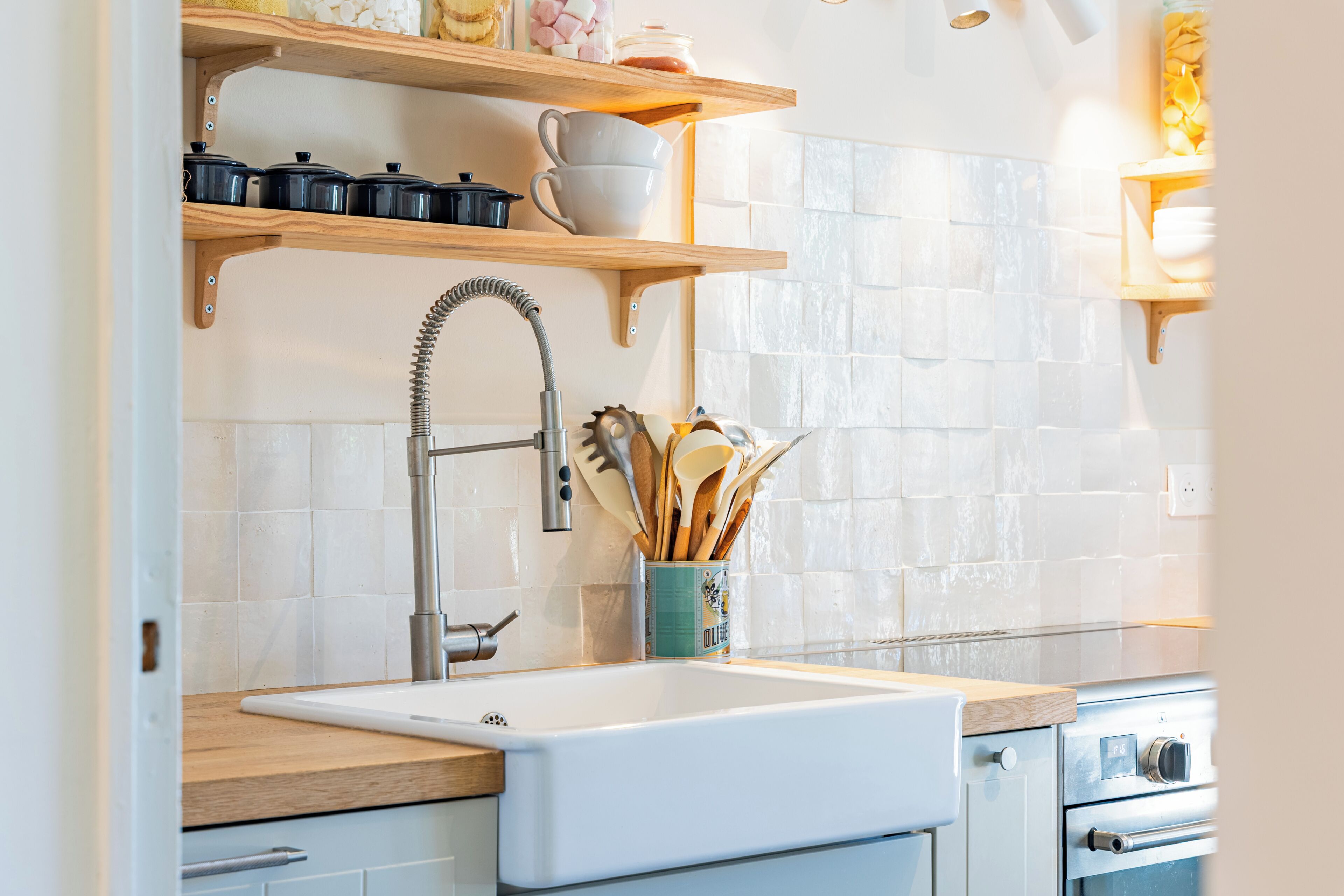 Une cuisine avec un évier blanc, des étagères en bois et un mur recouvert de carreaux.