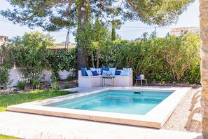 Et rektangulært swimmingpool med en hvid kant, omgivet af en betont terrasse. Der er et siddeområde med en hvid sofa og blå puder, samt et lille bord med en dekorativ genstand.