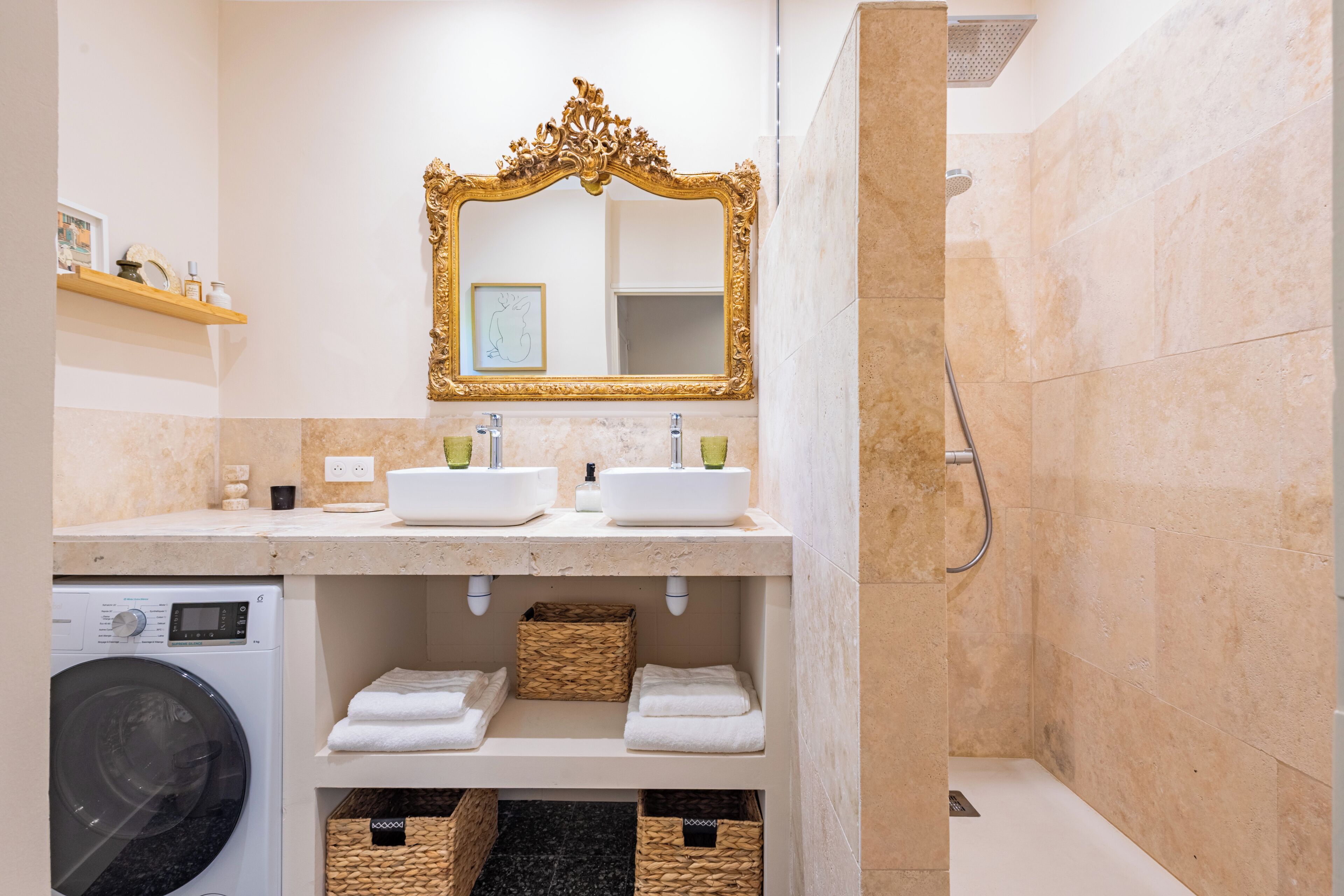 A bathroom with two sinks, a gold-framed mirror, a shower, and a washing machine.