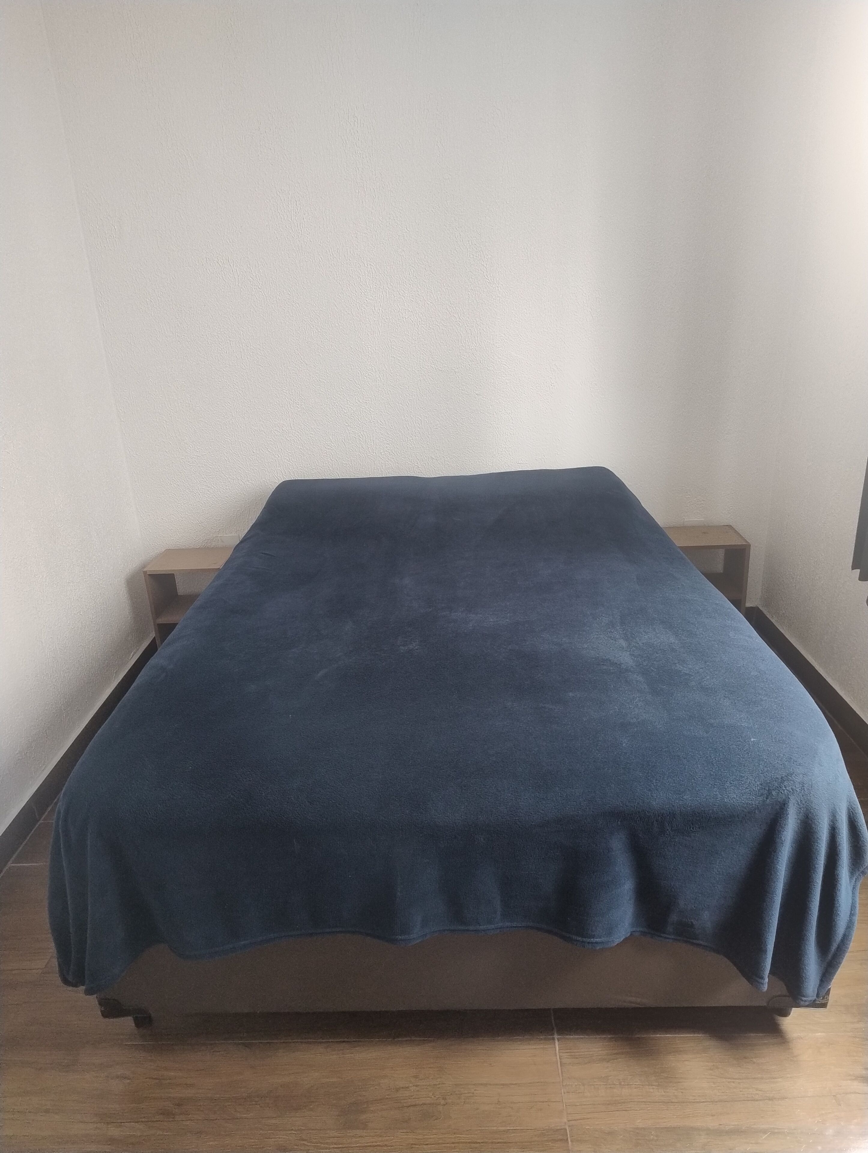 A single bed with a dark blue blanket in a room with plain walls and a wooden floor.