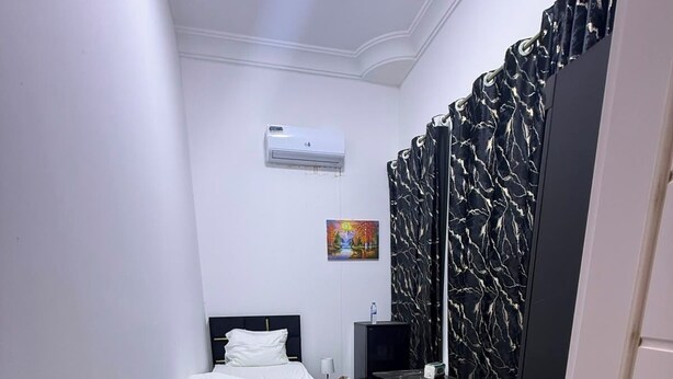 A small, narrow hotel room with a bed, a bedside table with a flower, a TV, and a black curtain with a leaf pattern. - Four Stars Residence (Dubai)