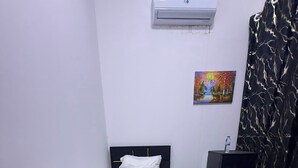 A small, single-bed room with a bed, bedside table, and a desk with a chair. There is a painting on the wall, a water bottle on the desk, and an air conditioning unit above the bed. - Four Stars Residence (Dubai)