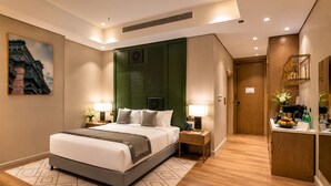 A hotel room with a large bed, bedside tables with lamps, a green accent wall, a painting on the wall, and a small kitchenette area.