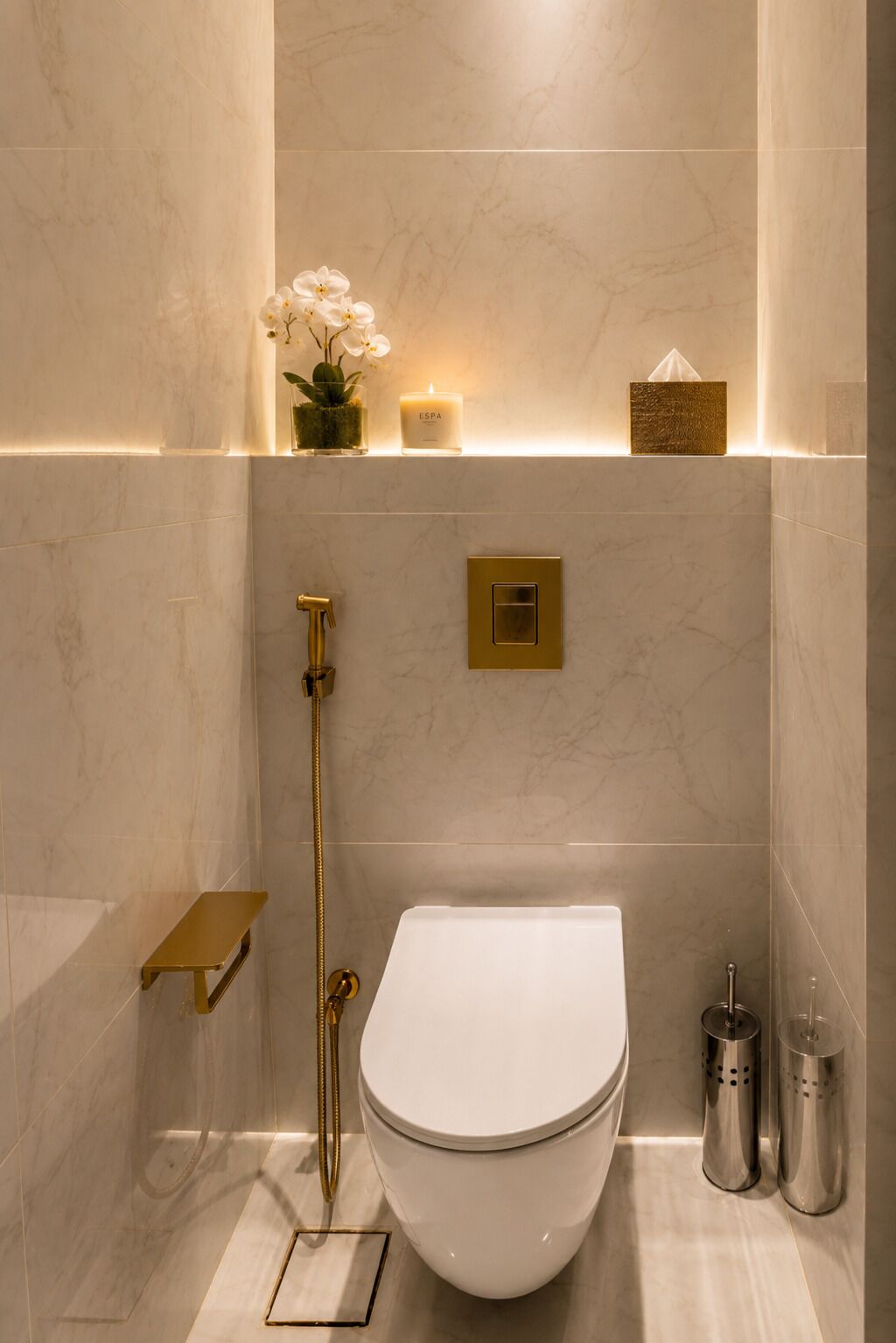 Une salle de bain moderne avec des toilettes blanches, une pomme de douche dorée et une petite étagère ornée d’une plante et d’une bougie.