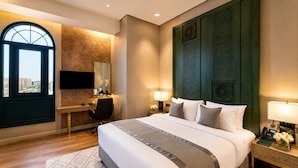 A hotel room with a large bed, a desk, a chair, a TV, and a view of the city. - Naviti By Warwick Jeddah (Al Quwaiiyah)