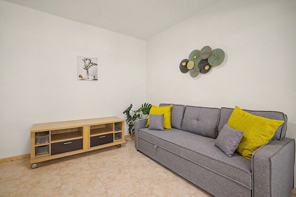 Ein Wohnzimmer mit grauem Sofa, gelben Kissen, einer Holzkombination mit Schubladen, einer Wanddekoration und einem Topfpflanzen.