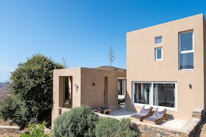 A modern two-story building with a flat roof, large windows, and a patio area with lounge chairs. - The Aer Element Of Gaea (Loutra)
