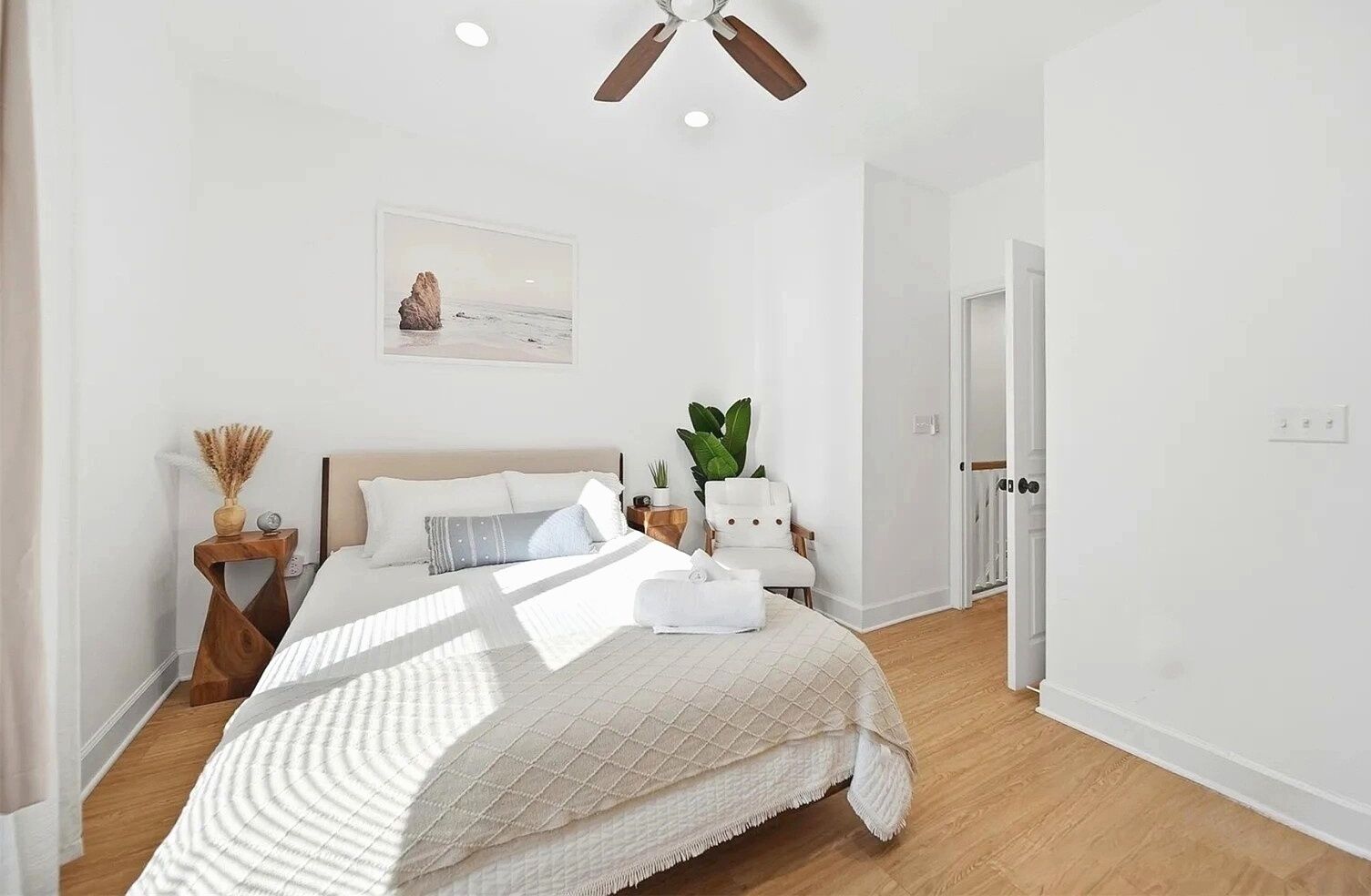 Une chambre à coucher comprenant un lit, des tables de chevet, un ventilateur de plafond, un tableau accroché au mur et une porte donnant sur une autre pièce.