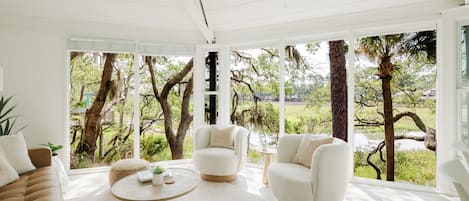 Una habitación amplia con ventanas grandes, una mesa de centro redonda, dos sillones blancos y un sofá marrón.