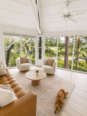 A spacious living room with a large window, a ceiling fan, a brown leather sofa, two armchairs, and a coffee table. - Canopy House - Walk to Harbour Town - Marshfront Oasis (Hilton Head Island)