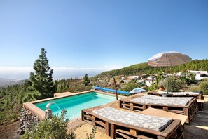 Una piscina con tumbonas de madera y una sombrilla, rodeada de vegetación y casas.