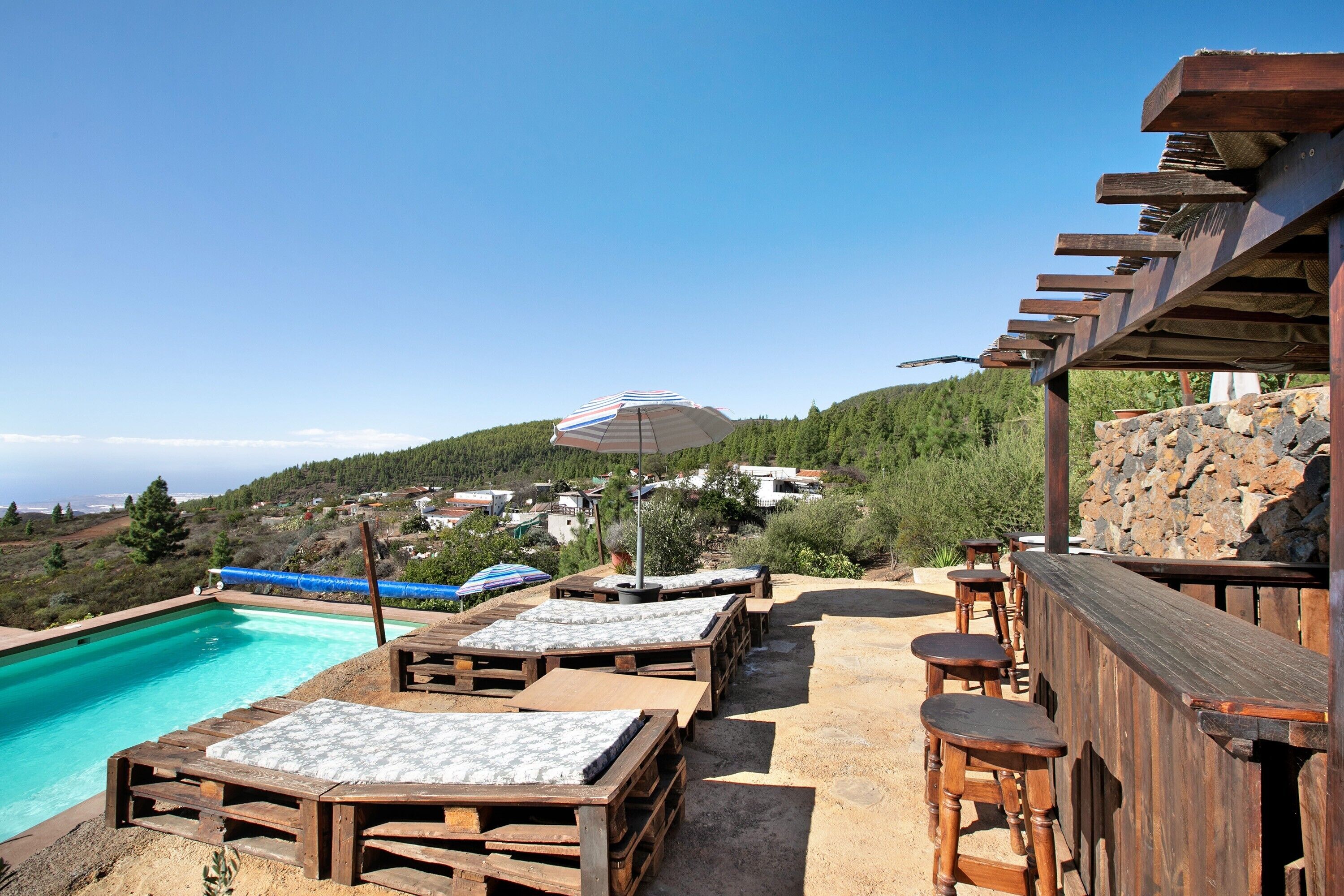 Ein Poolbereich mit Holzliegen und einer Bar mit Blick auf einen Hügel mit Häusern.