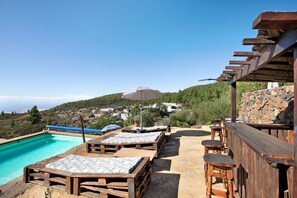 Ein Poolbereich mit Holzliegen und einer Bar mit Blick auf einen Hügel mit Häusern.