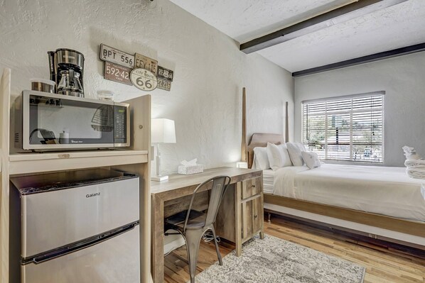 A bedroom with a bed, a desk with a chair, a microwave, a refrigerator, and a window with blinds.