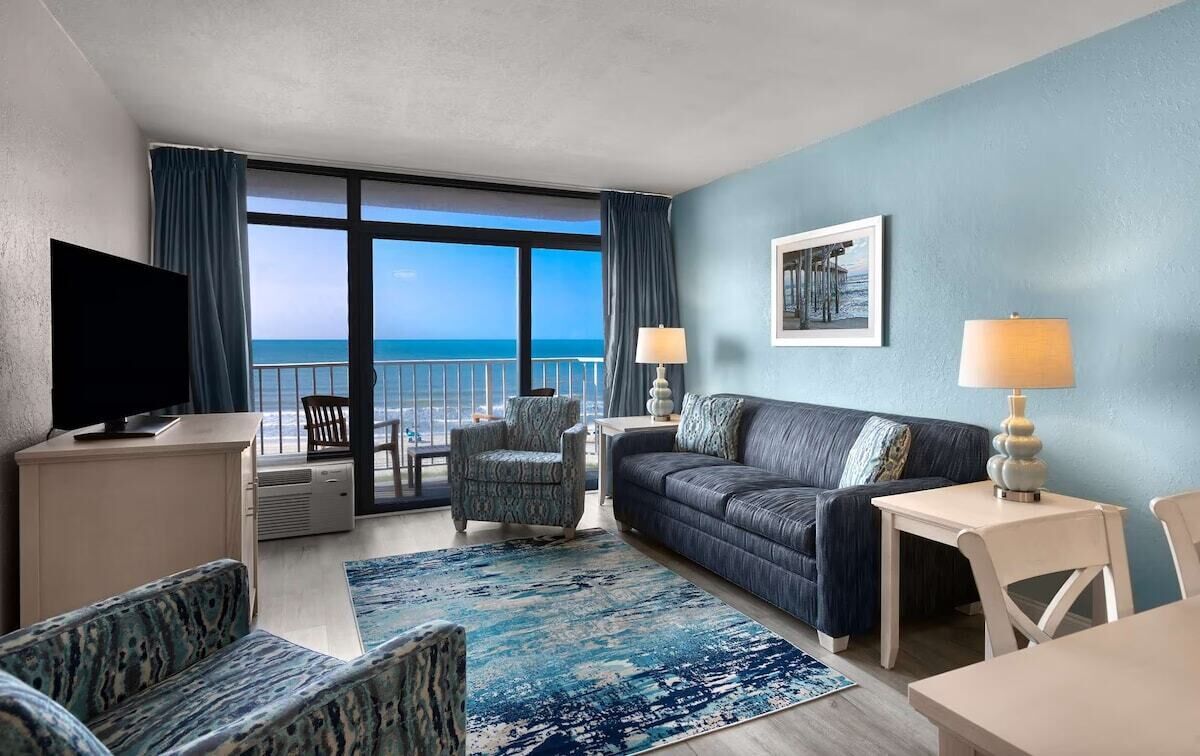A hotel room with a balcony, a sofa, a chair, a TV, and a table.
