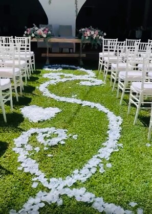 A venue with rows of white chairs arranged for an event, a central aisle with white petals on the grass, and floral arrangements on a table.