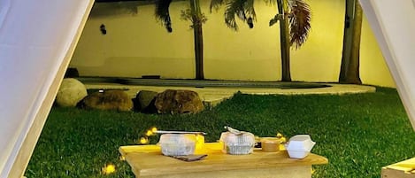 A tent set up on a grassy area with a table in front, surrounded by rocks and palm trees.