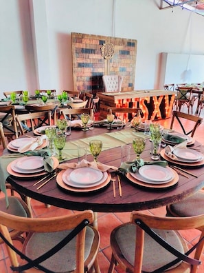 A dining area with round tables set for a meal, featuring wooden chairs and green glassware.