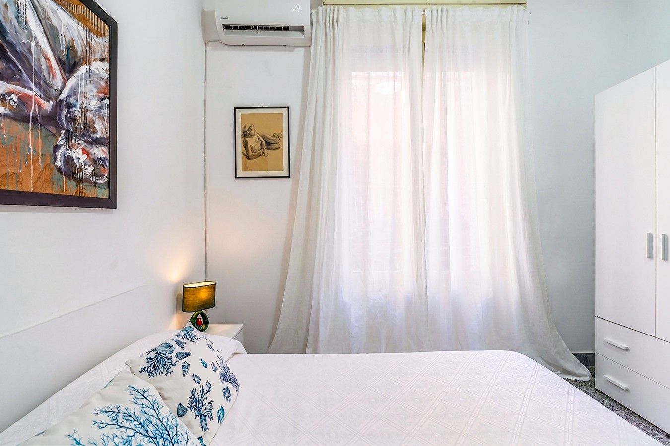 A bedroom with a bed, a nightstand, a lamp, a framed picture, a white wardrobe, and a window with sheer curtains.