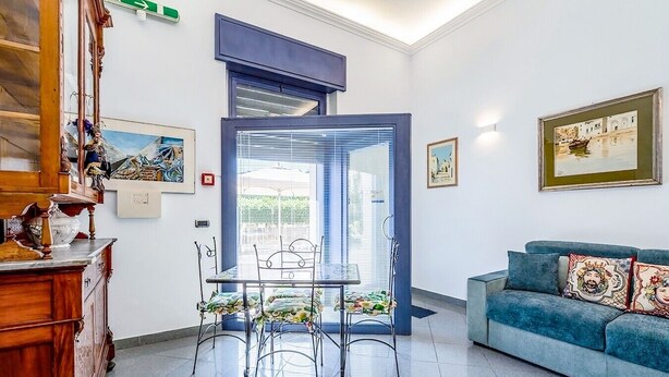 A living room with a blue sofa, a dining table with chairs, and a large window with blinds. - Al Portico - In the Heart of Avola (Avola)