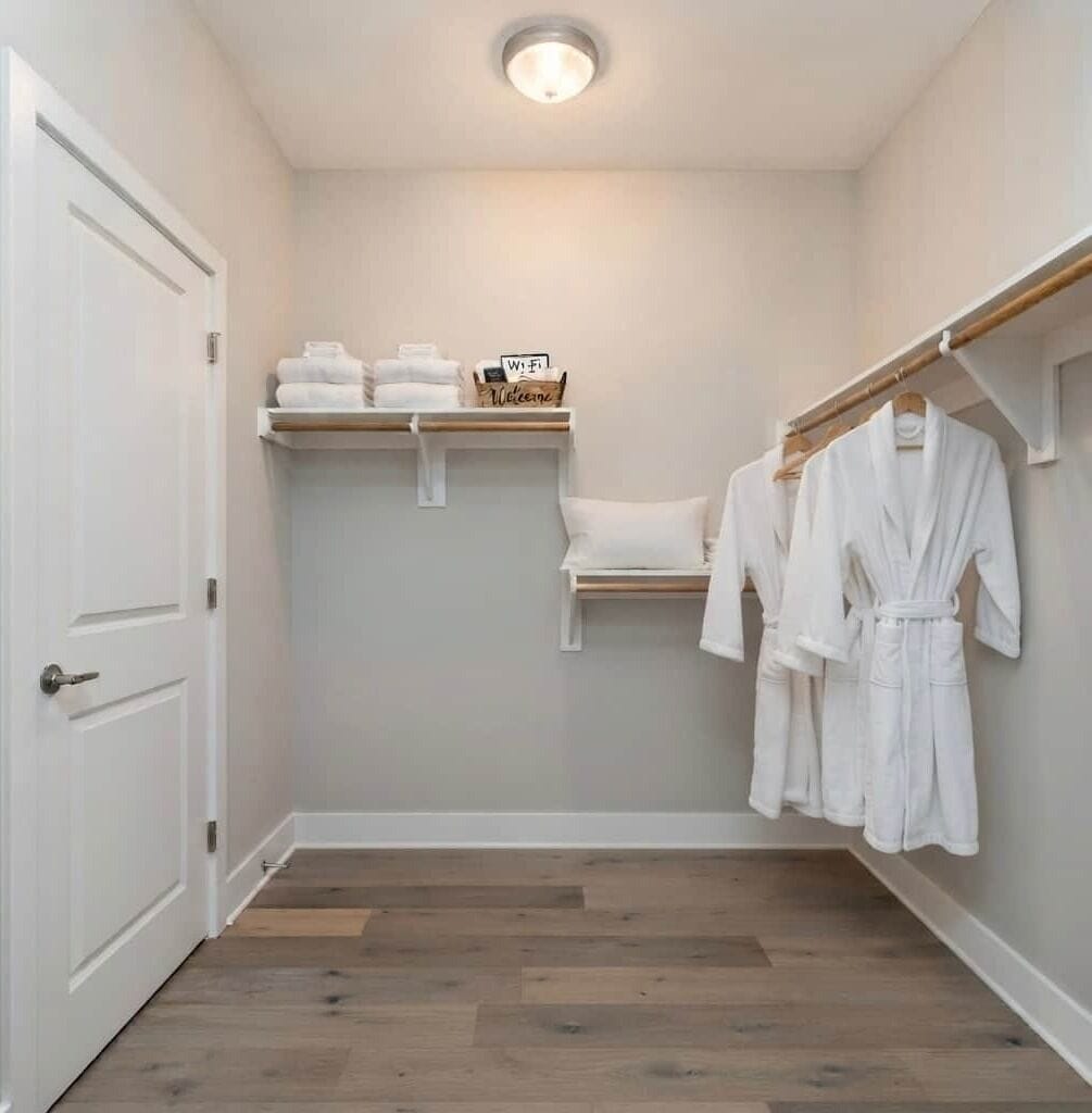Una habitación pequeña y bien iluminada con suelo de madera, una puerta blanca, un estante con toallas y un letrero, y un perchero con batas blancas.