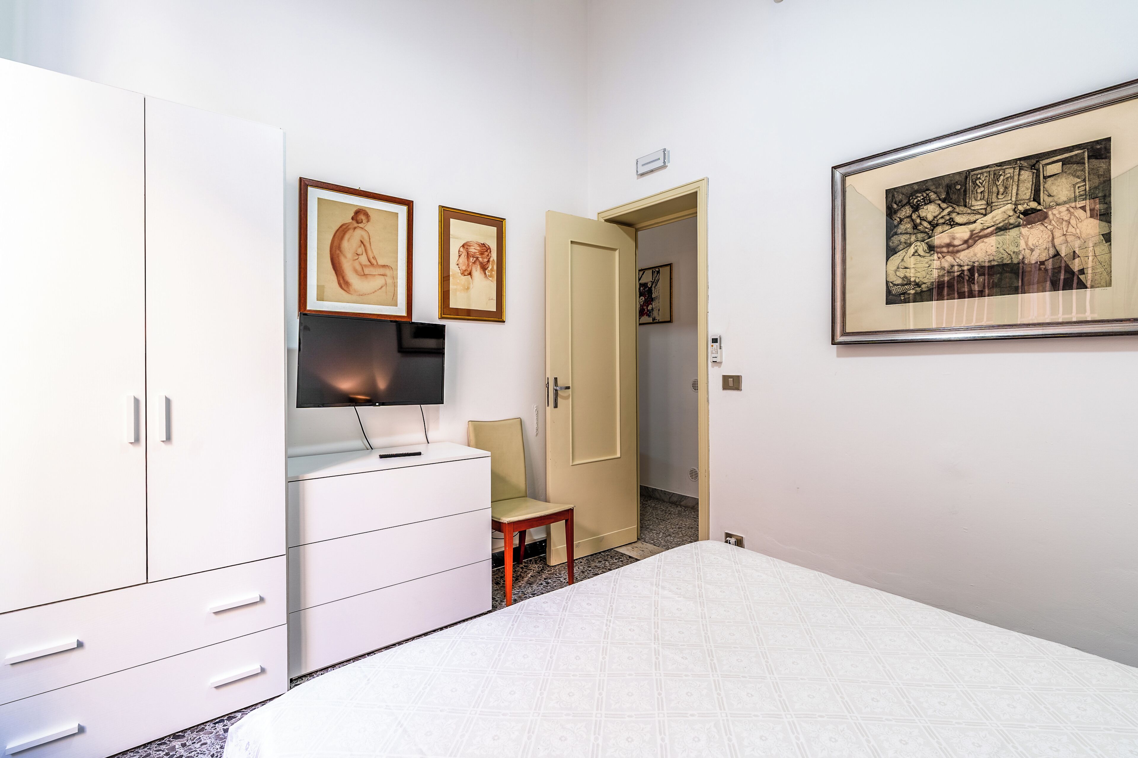A bedroom with a bed, a wardrobe, a desk with a chair, and artwork on the walls.