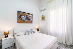 A hotel room with a bed, a nightstand, a wall-mounted air conditioner, and a window with curtains. - Al Portico (Avola)