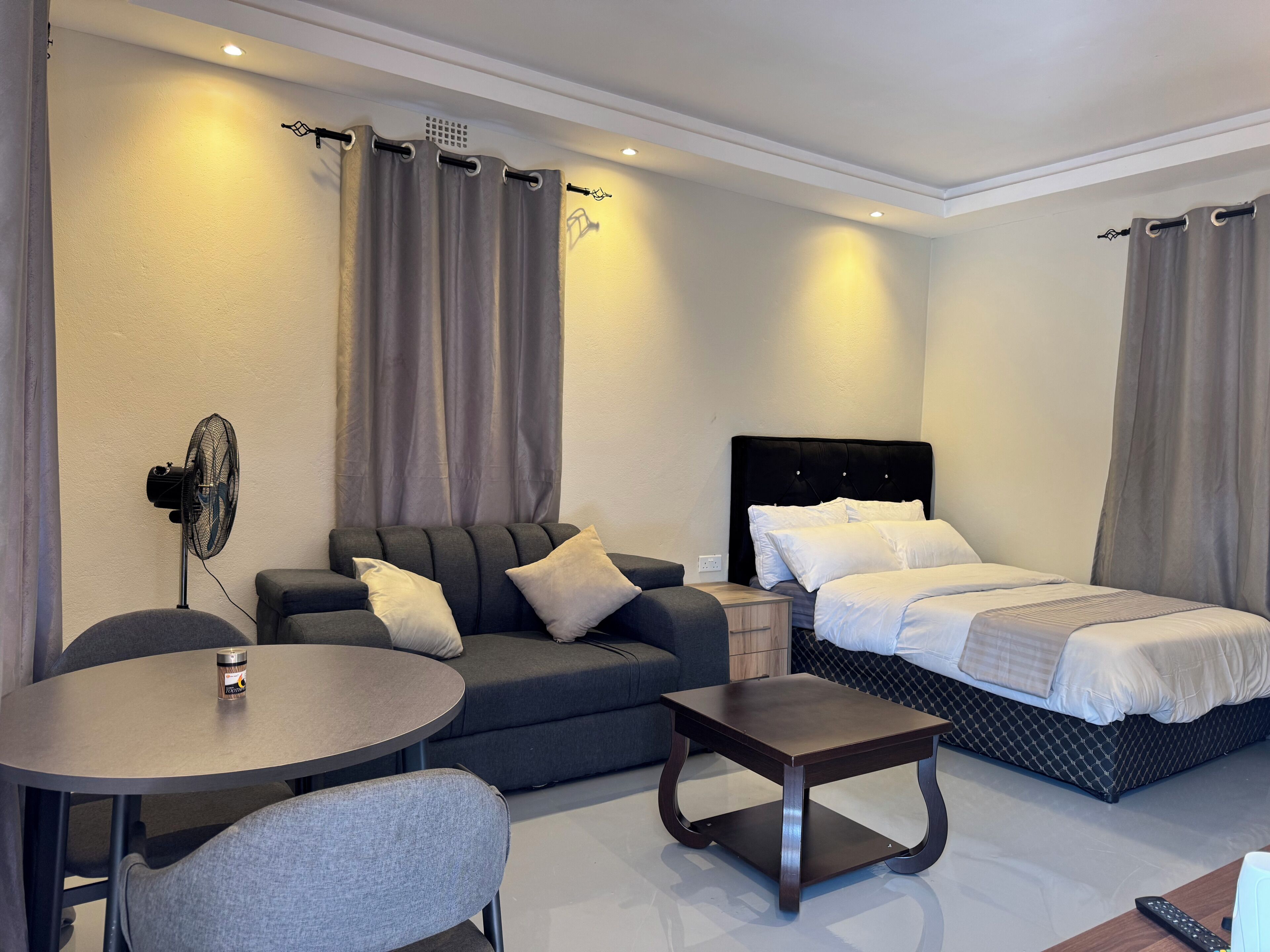 A hotel room with a bed, a sofa, a small table, a chair, and a standing fan.
